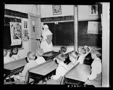 A black-and-white photograph of a Sister of the former Mont-Carmel convent school teaching a group of children at desks within a classroom. She points an instructional stick at a poster of a religious subject fixed to the classroom board while focusing her gaze on the children, ensuring they are attentive to her lesson.