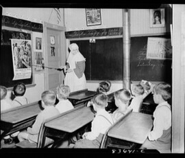 A black-and-white photograph of a Sister of the former Mont-Carmel convent school teaching a group of children at desks within a classroom. She points an instructional stick at a poster of a religious subject fixed to the classroom board while focusing her gaze on the children, ensuring they are attentive to her lesson.